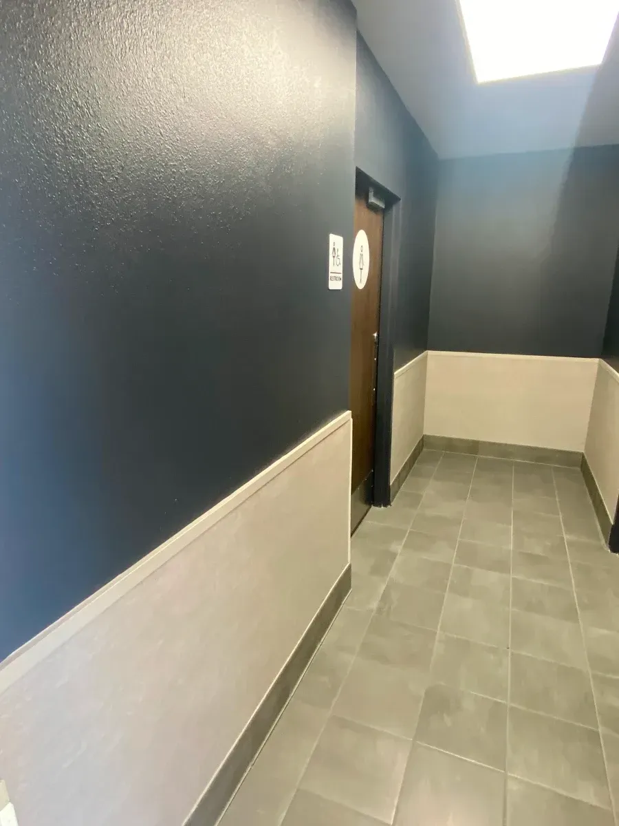 Narrow hallway with dark blue walls, beige wainscoting, and a restroom door.