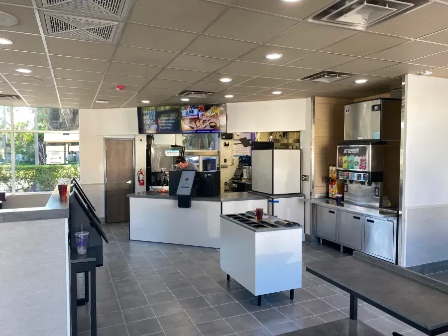 Interior of a fast-food restaurant; order counter, food prep area, drink dispenser, empty seating.