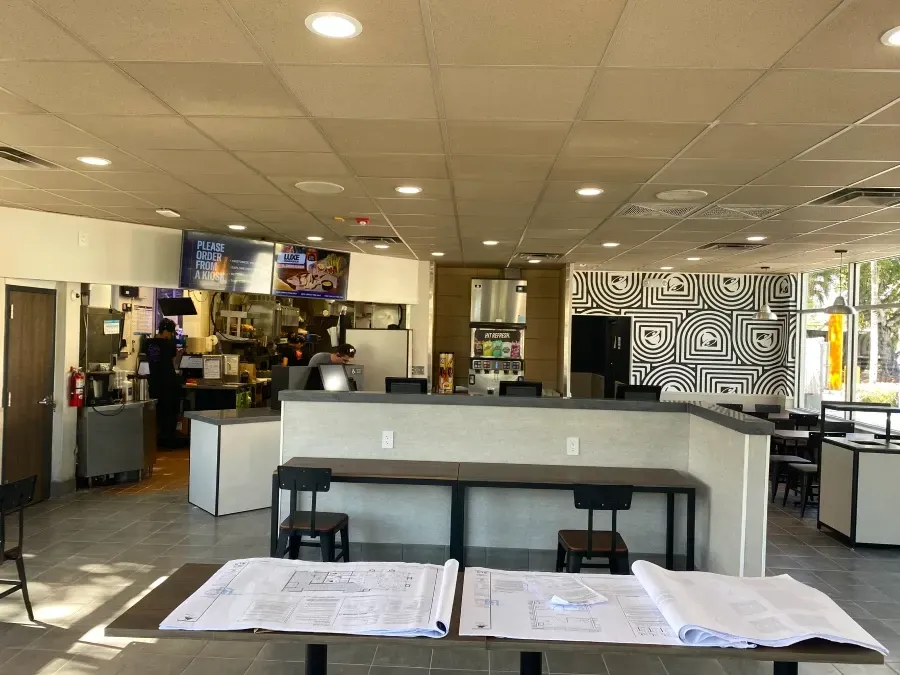 Interior of a restaurant under renovation. Architectural blueprints on a table in the foreground. Counter and seating areas visible.