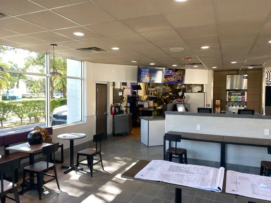 Interior of a fast-food restaurant with tables and chairs. Kitchen staff visible behind the counter.
