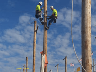 Two utility workers in neon vests and helmets work at the top of a wooden utility pole against a blue, cloudy sky.