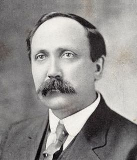 Black-and-white portrait of a man with a mustache, wearing a collared shirt, necktie, and suit jacket.