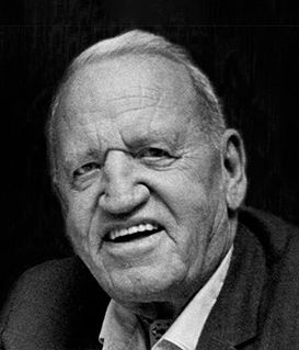 A black-and-white portrait of a smiling person wearing a collared shirt and jacket against a dark background.