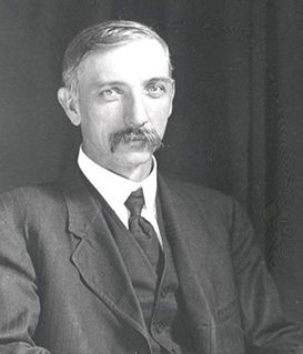 A black-and-white portrait of a man in a formal suit and necktie, posed against a dark, plain background.