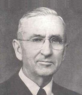 A black-and-white portrait of a person wearing round glasses, a collared shirt, and a suit jacket, looking at the camera.