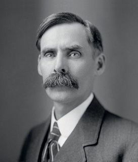 Portrait of a person with a mustache, wearing a collared shirt, tie, and suit jacket against a plain background.