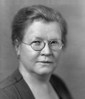 A studio portrait of a person wearing round glasses and a dark, high-collared garment against a plain backdrop.