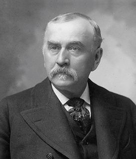 A formal black-and-white portrait of a man with a mustache, wearing a suit and cravat, against a mottled backdrop.
