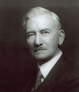 A black-and-white portrait of a man in a suit, tie, and mustache, facing forward with a neutral expression.