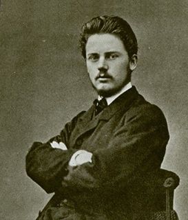 A sepia-toned portrait of a person with a mustache, wearing a dark suit, seated with arms crossed.