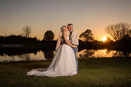 A bride and groom in wedding attire embrace at sunset by a calm lake with silhouettes of trees in the background.