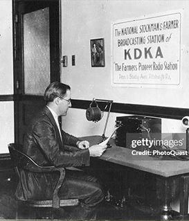 A person sits at a desk reading into a microphone at radio station KDKA, with a sign on the wall behind them.