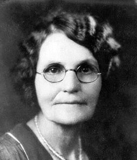 A black-and-white portrait of a person with wavy hair wearing round glasses and a collared garment with a beaded necklace.