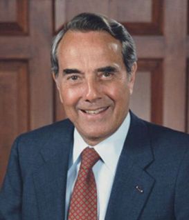 Bob Dole wearing a navy blue suit and red patterned tie, smiling in front of a wood-paneled wall.
