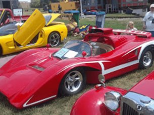 A bright red sports car with white stripes parked on grass at an outdoor car show, with a yellow car in the background.