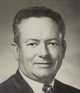 A black-and-white portrait of a man in a suit and tie, smiling slightly against a solid background.