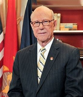 Official portrait of Congressman Don Young wearing a suit and tie, positioned in front of US and state flags and books.