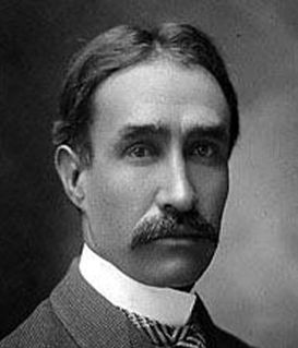 A studio portrait of a man with a mustache, wearing a collared shirt, necktie, and jacket.