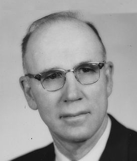 A formal portrait of a person wearing glasses and a suit, looking toward the camera against a plain background.