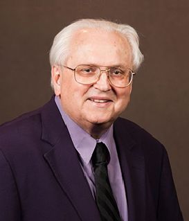 A person wearing glasses, a dark blazer, a light purple shirt, and a dark tie, smiling against a solid brown background.
