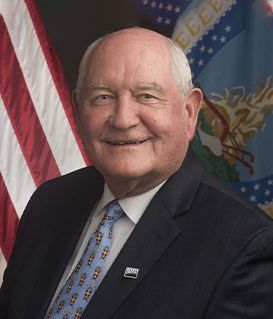 Sonny Perdue smiling in a suit and tie, posed in front of the American flag and the USDA departmental flag.