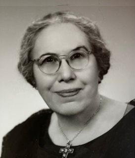 A portrait of a person with wavy hair and glasses, wearing a cross necklace against a light background.