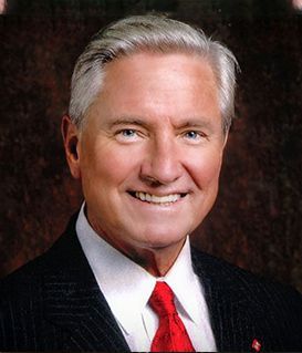A smiling person with grey hair, wearing a dark pinstripe blazer, white collared shirt, and a red tie against a dark wall.