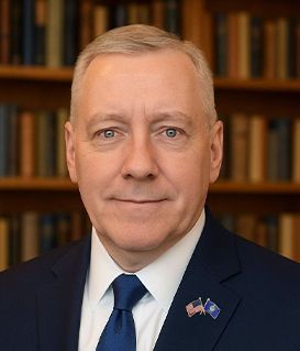 A man in a suit and blue tie stands before a bookshelf. He wears an American flag and a state flag lapel pin.