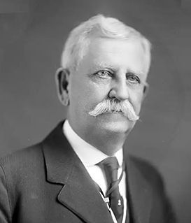 A head-and-shoulders studio portrait of a man with a white handlebar mustache, wearing a suit and tie.