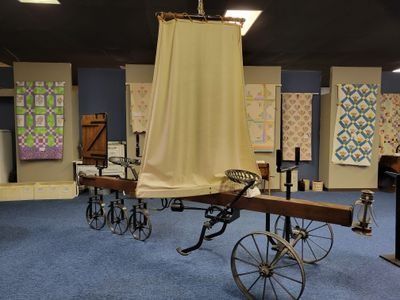 An antique agricultural implement with a tall, tan canvas screen on display in a museum room with wall-mounted quilts.