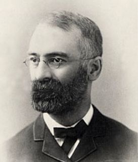 A black-and-white portrait of a man with a beard and glasses, wearing a suit and bowtie against a plain background.