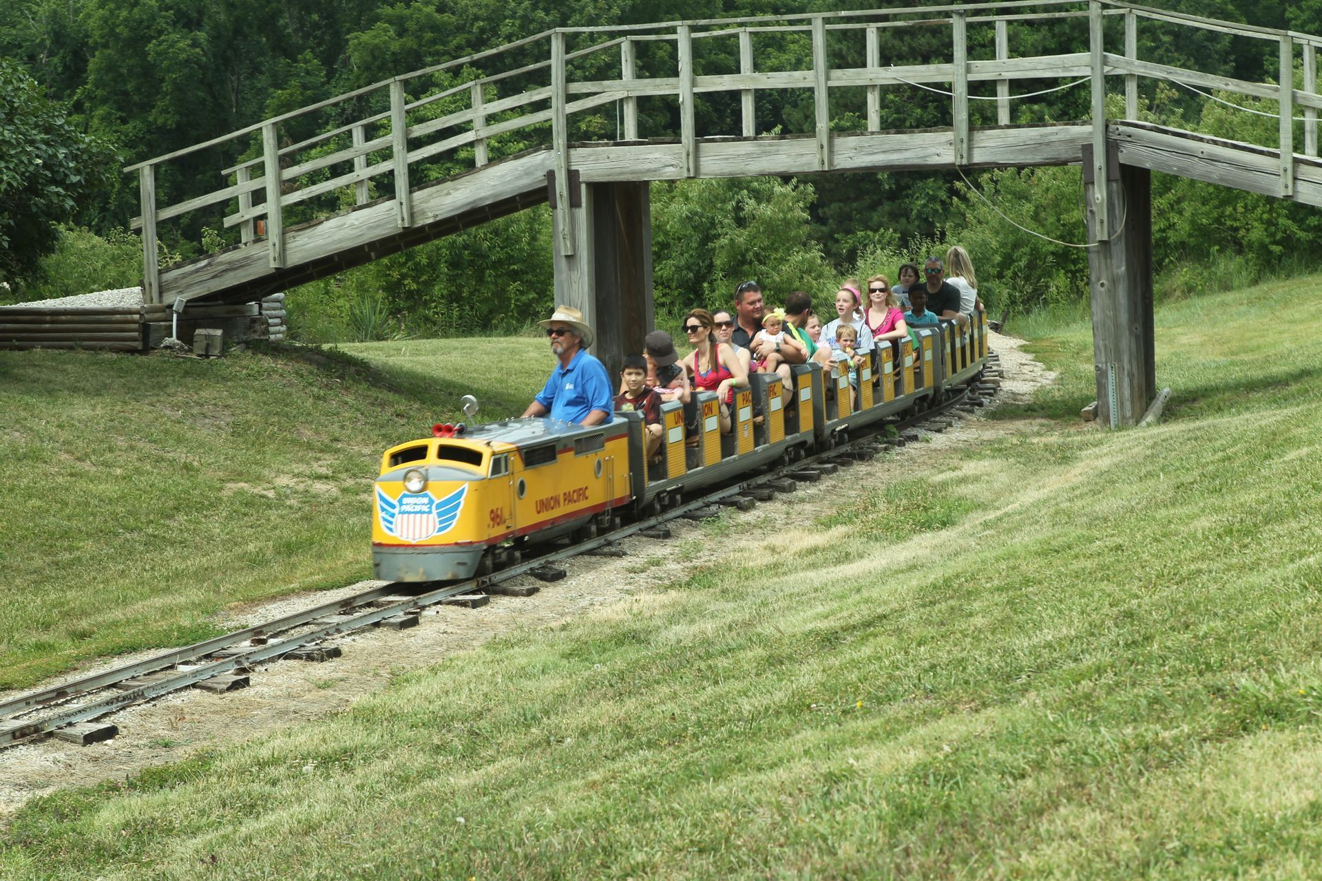A yellow miniature train with passengers riding along tracks beneath a wooden footbridge in a grassy field.