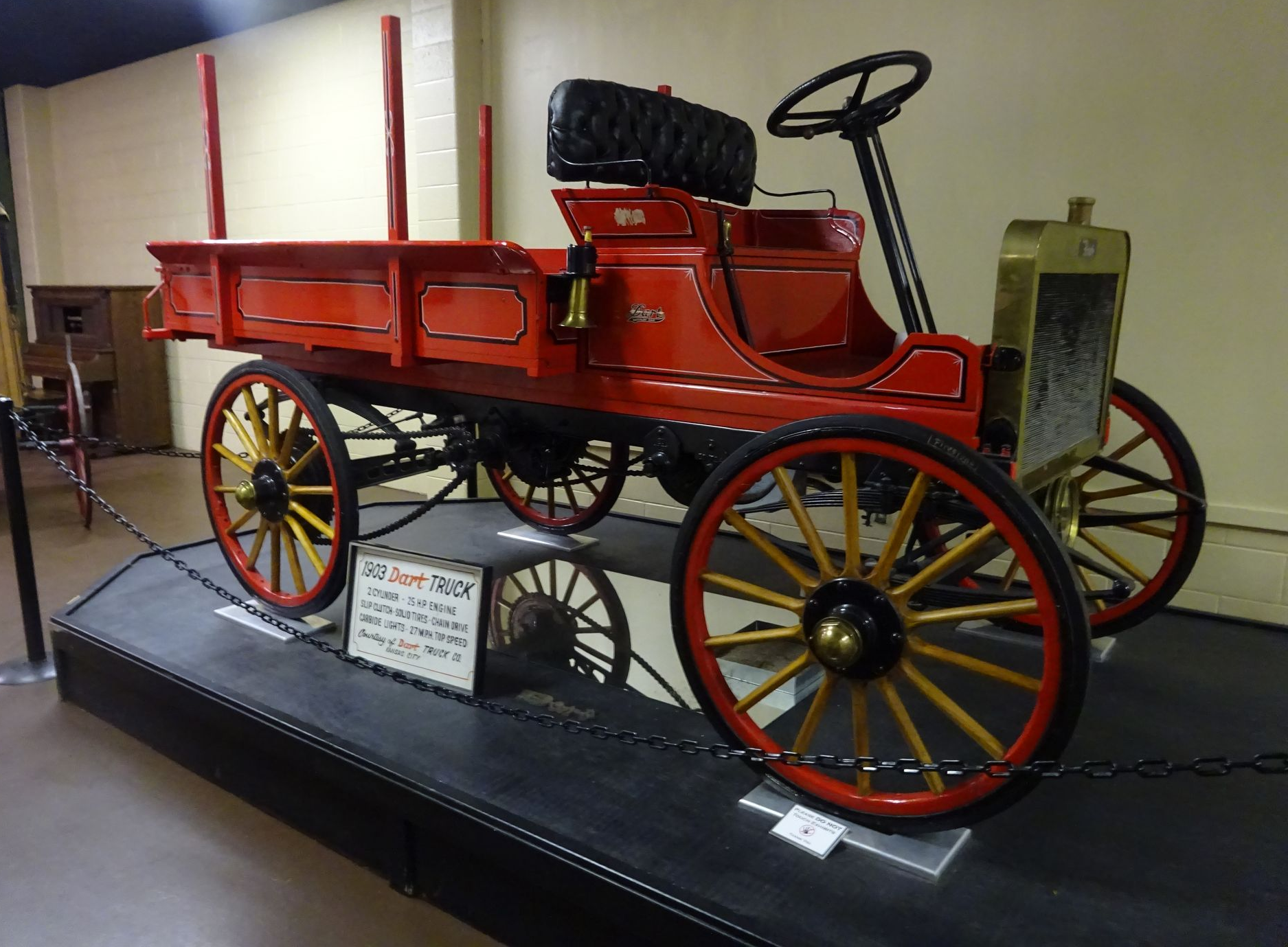 A vintage red wooden flatbed truck with large spoked wheels displayed on a raised platform in an indoor museum.