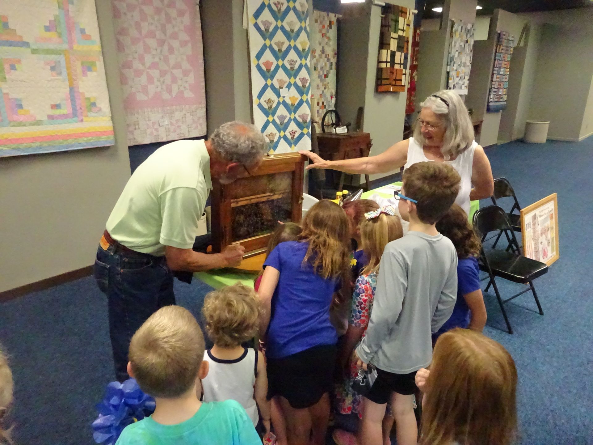 A man and woman show a small, box-like exhibit to a group of children in a gallery decorated with hanging quilts.