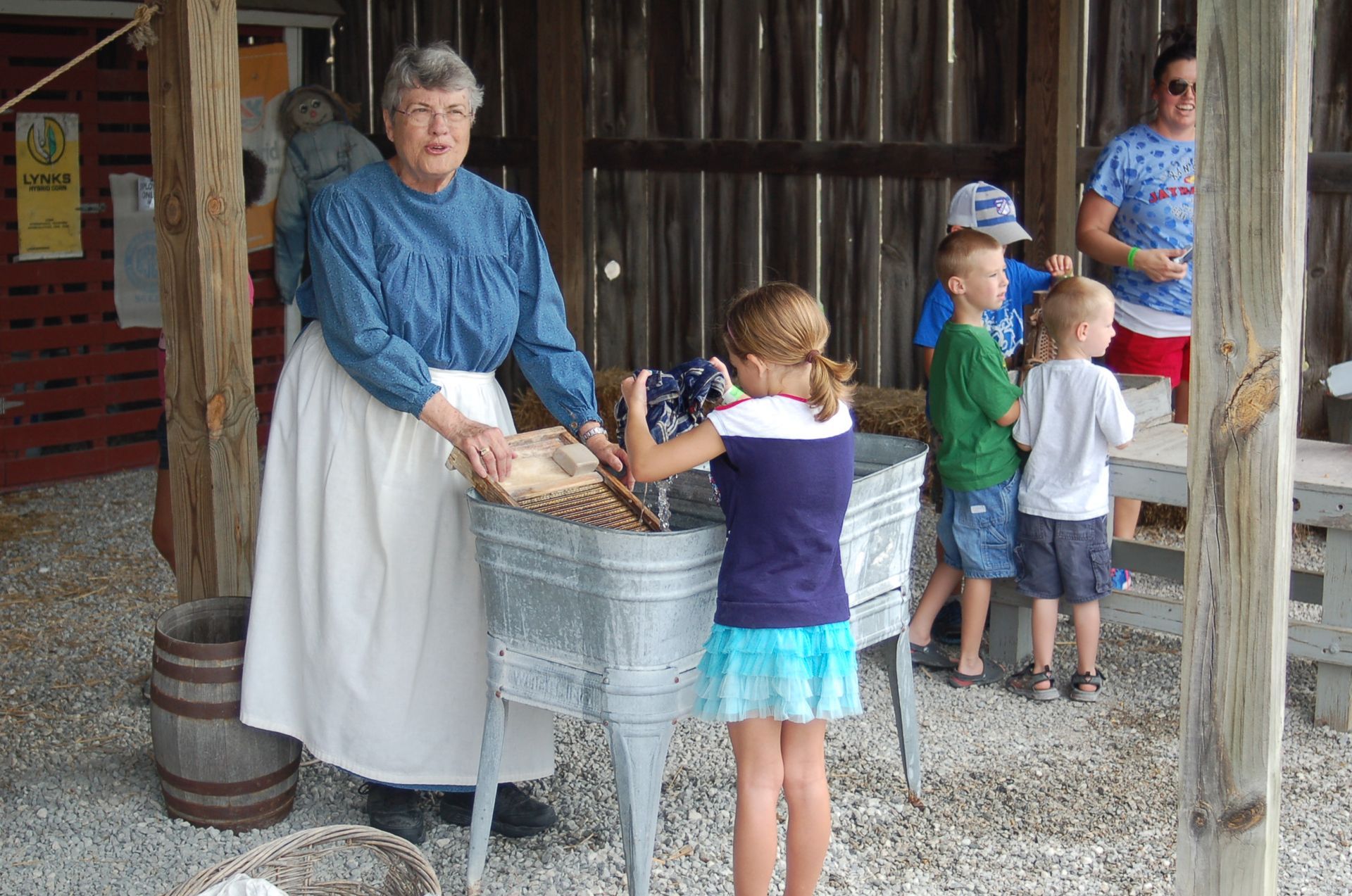 A person in historical clothing teaches a child how to use a scrub board in a metal washbasin in an open-air barn.