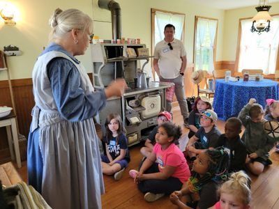 A costumed historical interpreter speaks to a seated group of children in a room with a vintage wood-burning stove.