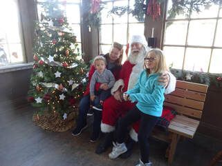 Santa Claus sitting on a bench with two children by a decorated Christmas tree in a sunlit room.