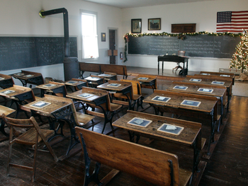 A historic one-room schoolhouse interior featuring rows of vintage wooden desks, a blackboard, and an American flag.
