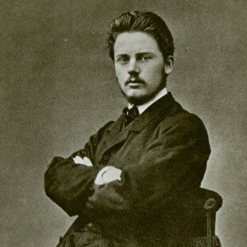 A vintage, sepia-toned portrait of a person with dark hair and a mustache, wearing a dark suit, sitting with arms crossed.