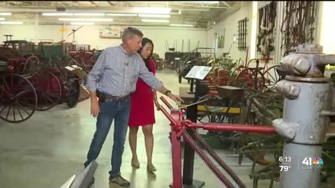 A man in a light blue shirt and a woman in a red dress stand in a museum, looking at a red antique farm tool.