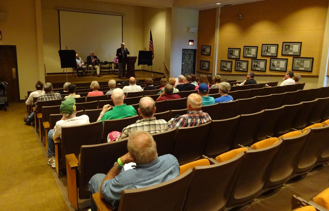 A speaker stands at a podium in a meeting hall, addressing an audience seated in rows of chairs facing the front stage.
