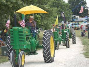A line of green John Deere tractors with yellow umbrellas and American flags traveling along a dirt path in a park.