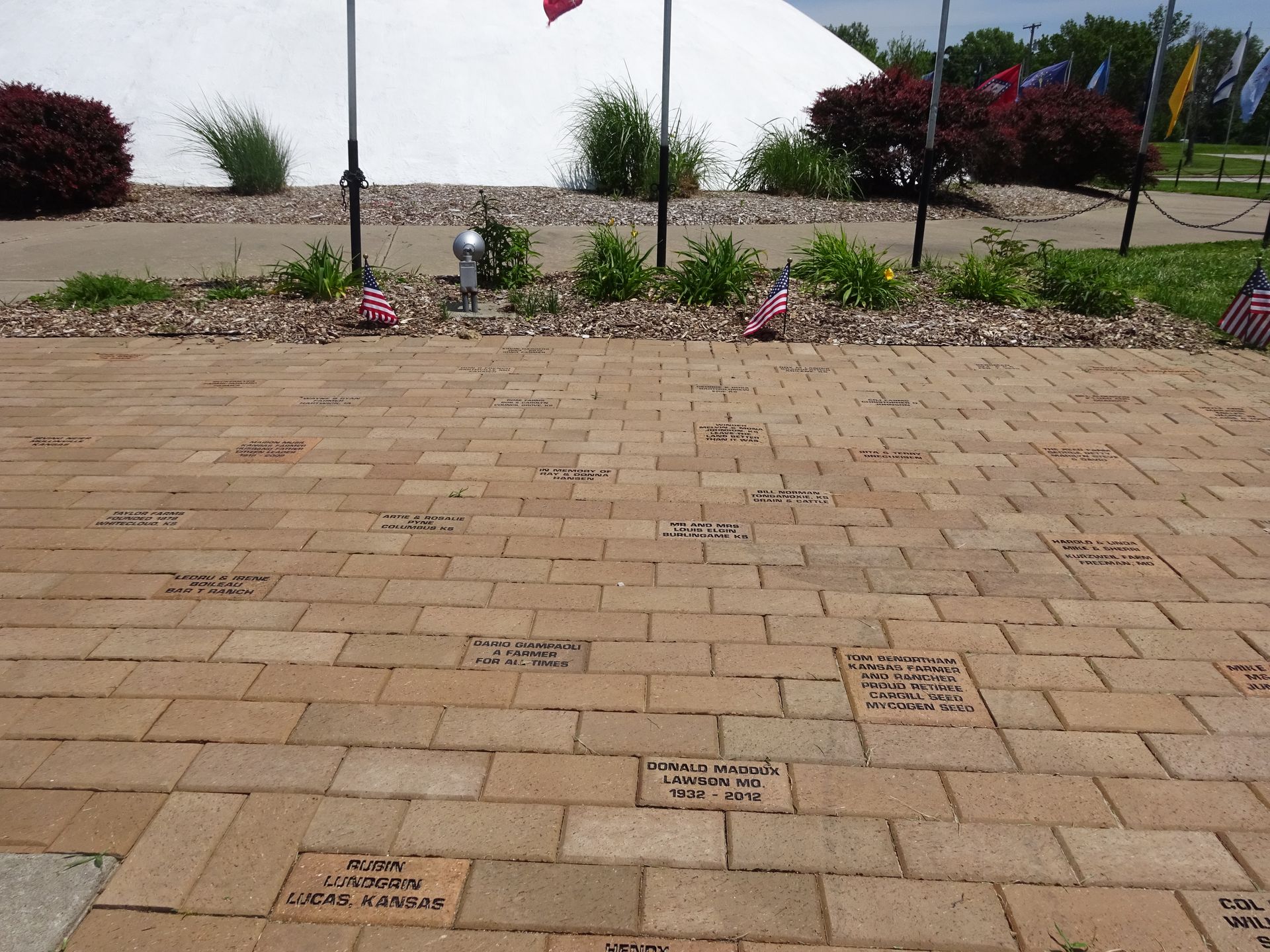 A paved walkway filled with memorial bricks inscribed with names, set in front of a garden with flags and greenery.