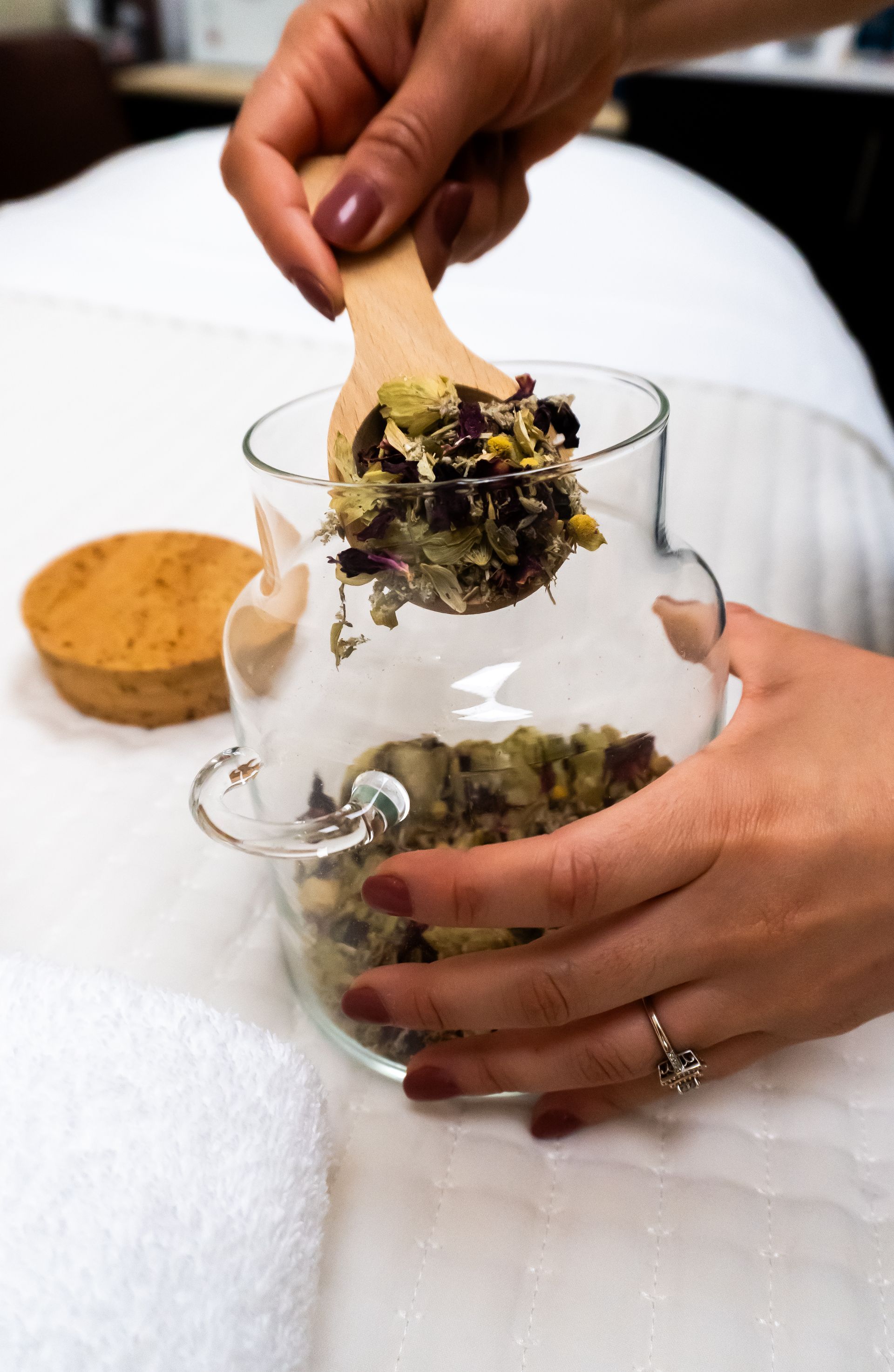 Hands pouring herbs from a wooden spoon into a glass jar, next to a cork.