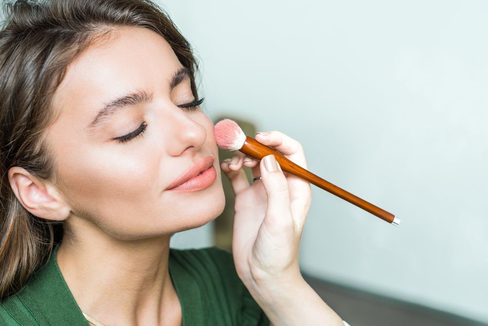 Woman applying blush with a brush to her cheek, eyes closed.