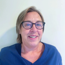 Kvinne med briller, smilende, iført blå skrubber, innendørs med en hvit veggbakgrunn.