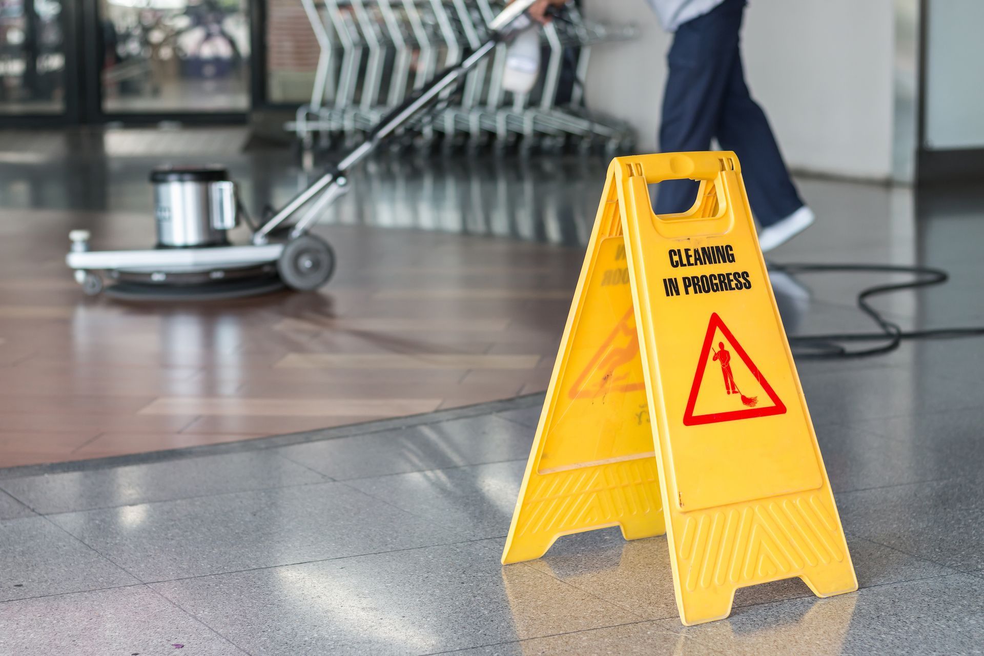 Yellow “Cleaning in Progress” sign on floor with cleaner using buffer in background. Yellow “Cleaning in Progress” sign on floor with cleaner using buffer in background.