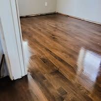Newly refinished hardwood floors in a room, reflecting light. White door frame in the left corner.