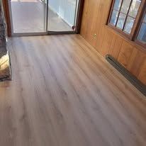 Light wood-look flooring fills a room; a sliding glass door and a wooden wall are visible.