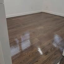 Shiny, light brown hardwood floor in a room, reflecting light. White wall in the background.